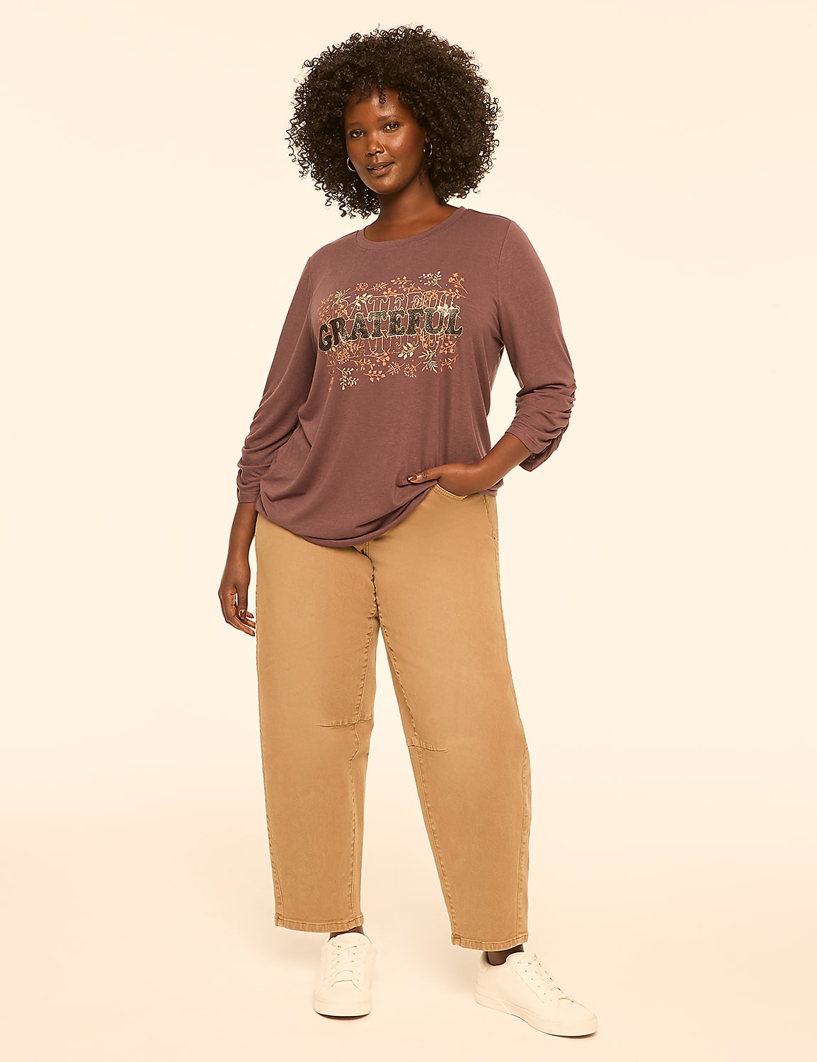 A woman with dark curly hair stands in the foreground wearing a long-sleeved shirt with the word "GRATEFUL" and floral accents, and loose-fitting tan pants, against a plain, light-colored background. Product Image 3