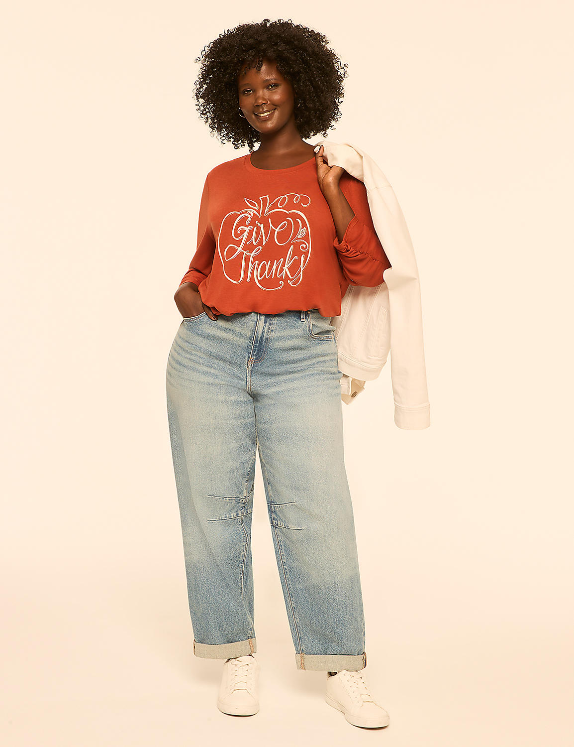 A woman in a rust-colored sweater and light-wash jeans stands against a plain, peachy background, holding a white jacket over her shoulder. Product Image 3