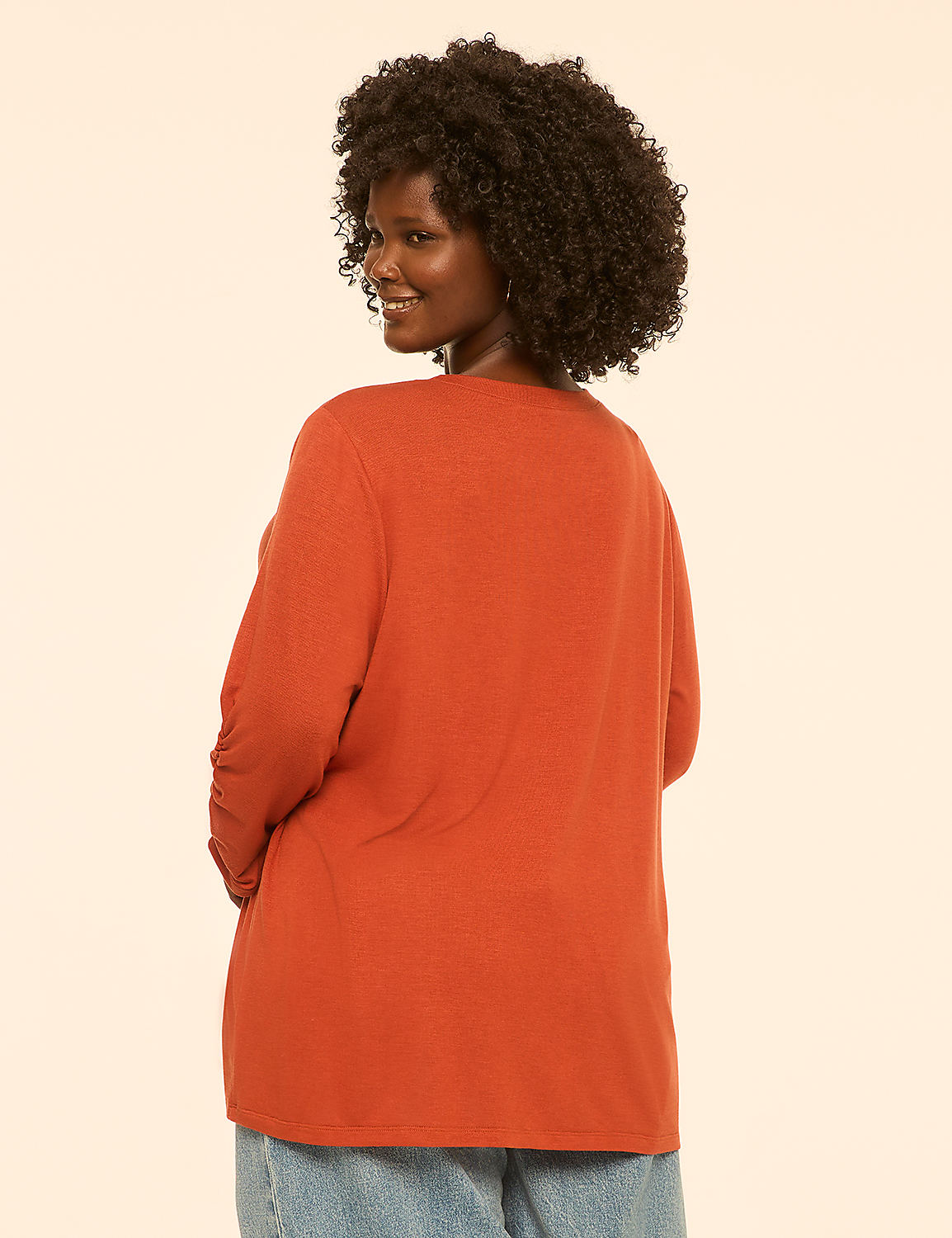A woman with curly dark hair, wearing an orange long-sleeved shirt and blue jeans, stands against a plain, light-colored background. Product Image 2