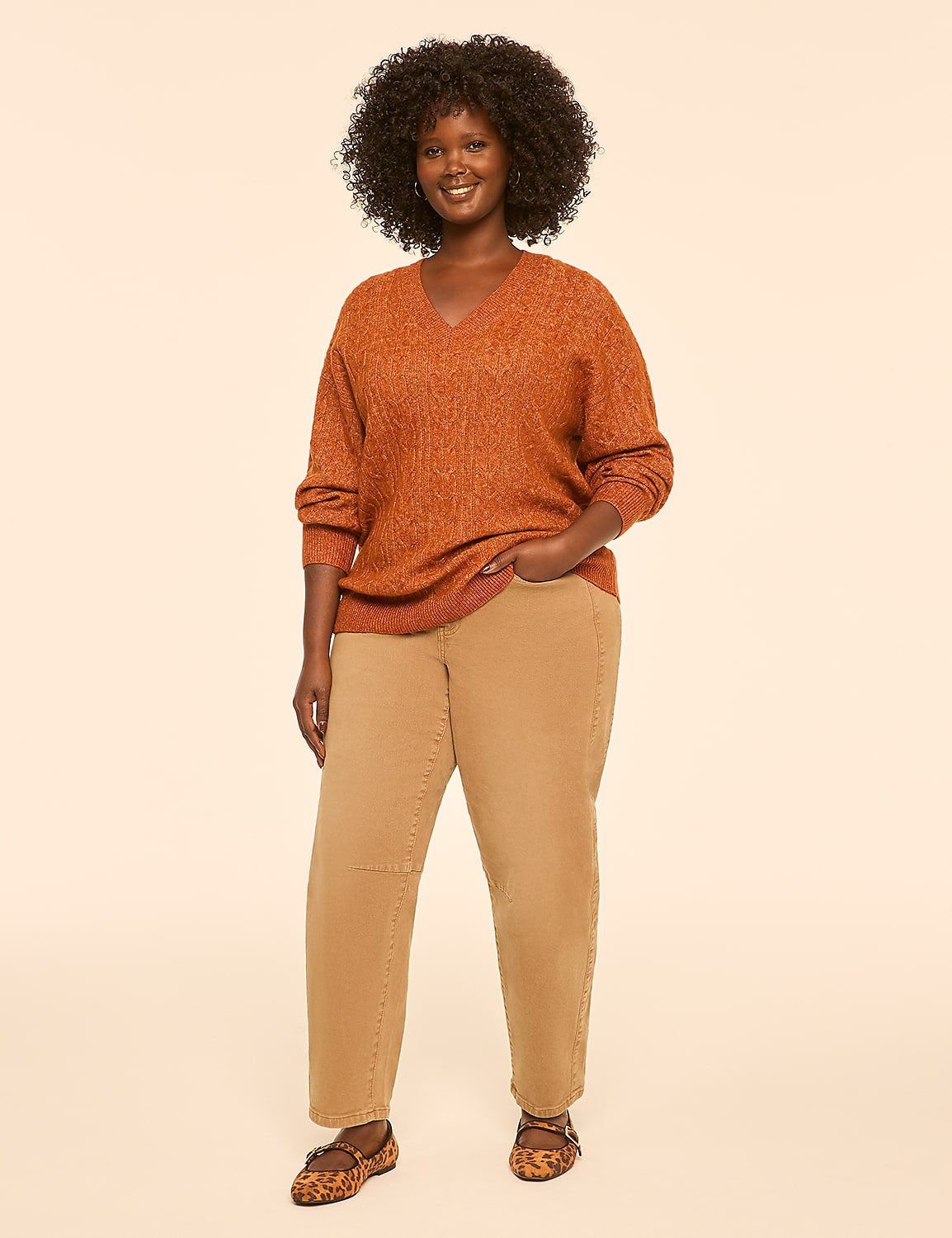 A woman with dark, curly hair stands in the foreground wearing an orange sweater and tan pants, against a plain, light-colored background. Product Image 3