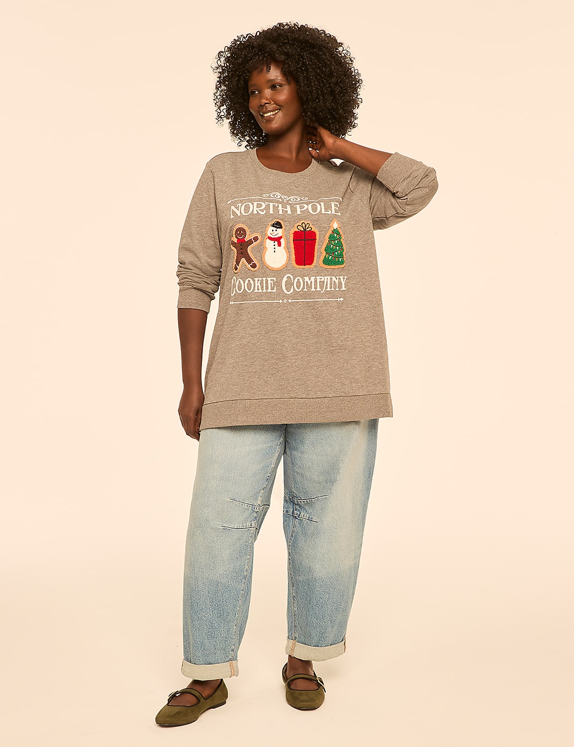 A woman in a grey sweatshirt with a Christmas cookie design and light-wash jeans stands against a plain, light-colored background. Product Image 3