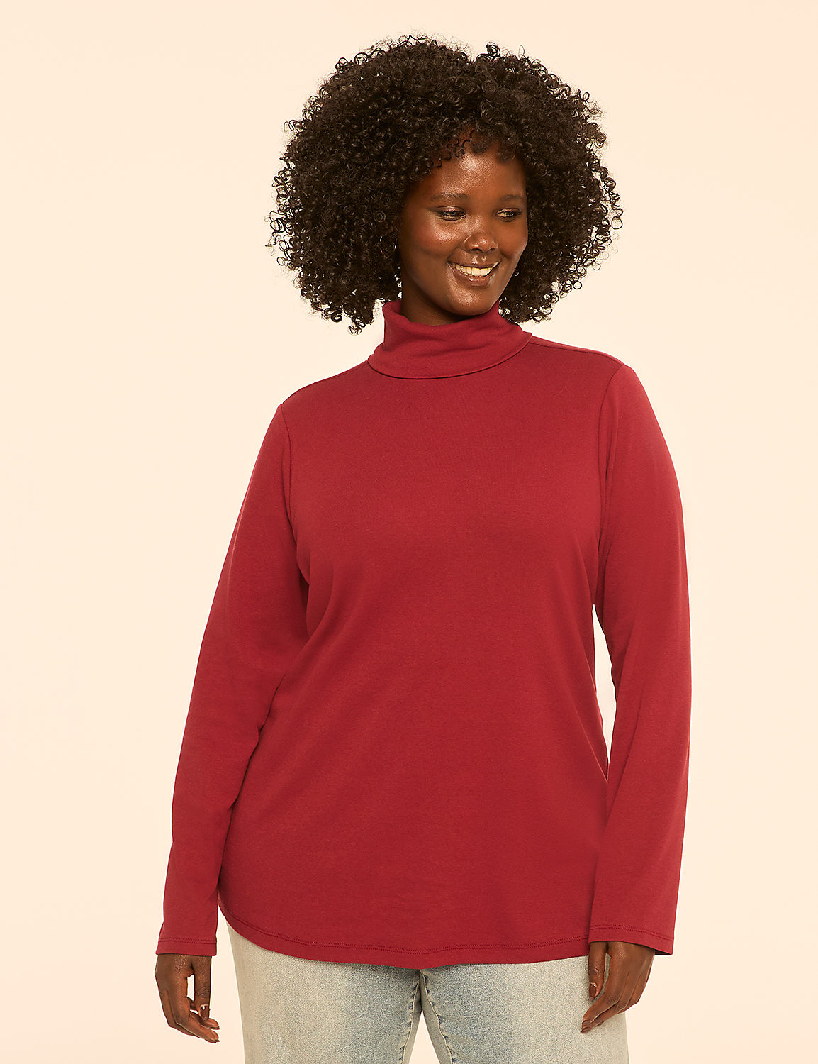 A smiling woman with curly dark hair wears a maroon turtleneck and light blue jeans against a plain cream background. Product Image 1