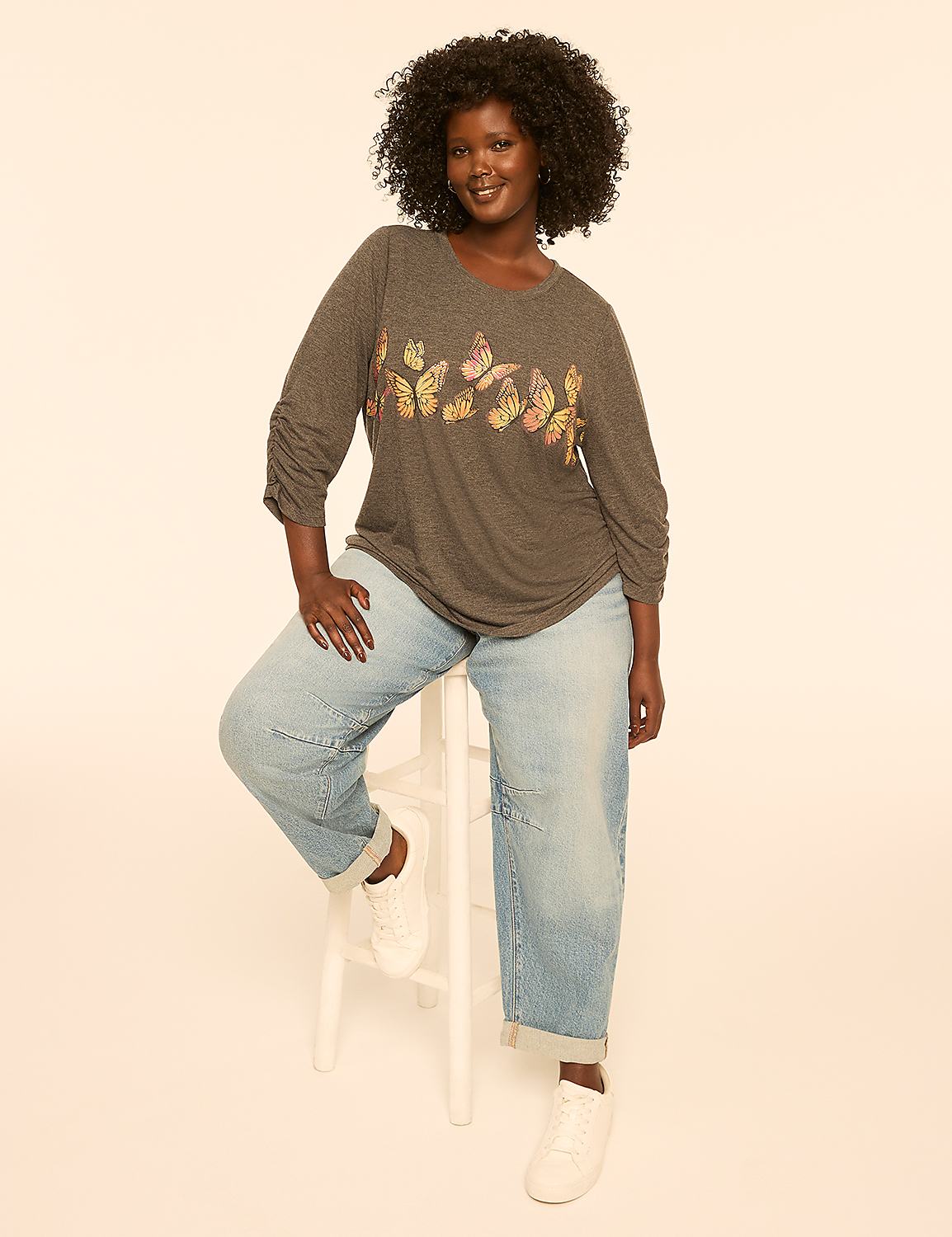 A woman with dark curly hair sits on a white stool wearing a gray t-shirt with butterflies and light-wash jeans, against a plain, light-colored background. Product Image 3
