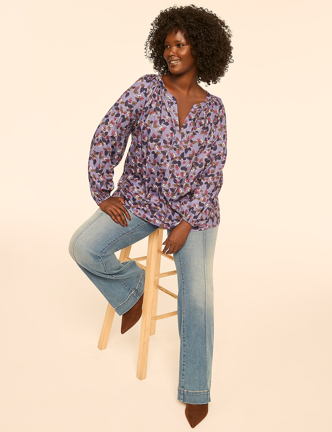A woman with dark curly hair sits on a wooden stool in the foreground, wearing a patterned purple blouse and light-wash jeans, against a plain, light-colored background. Product Image 3