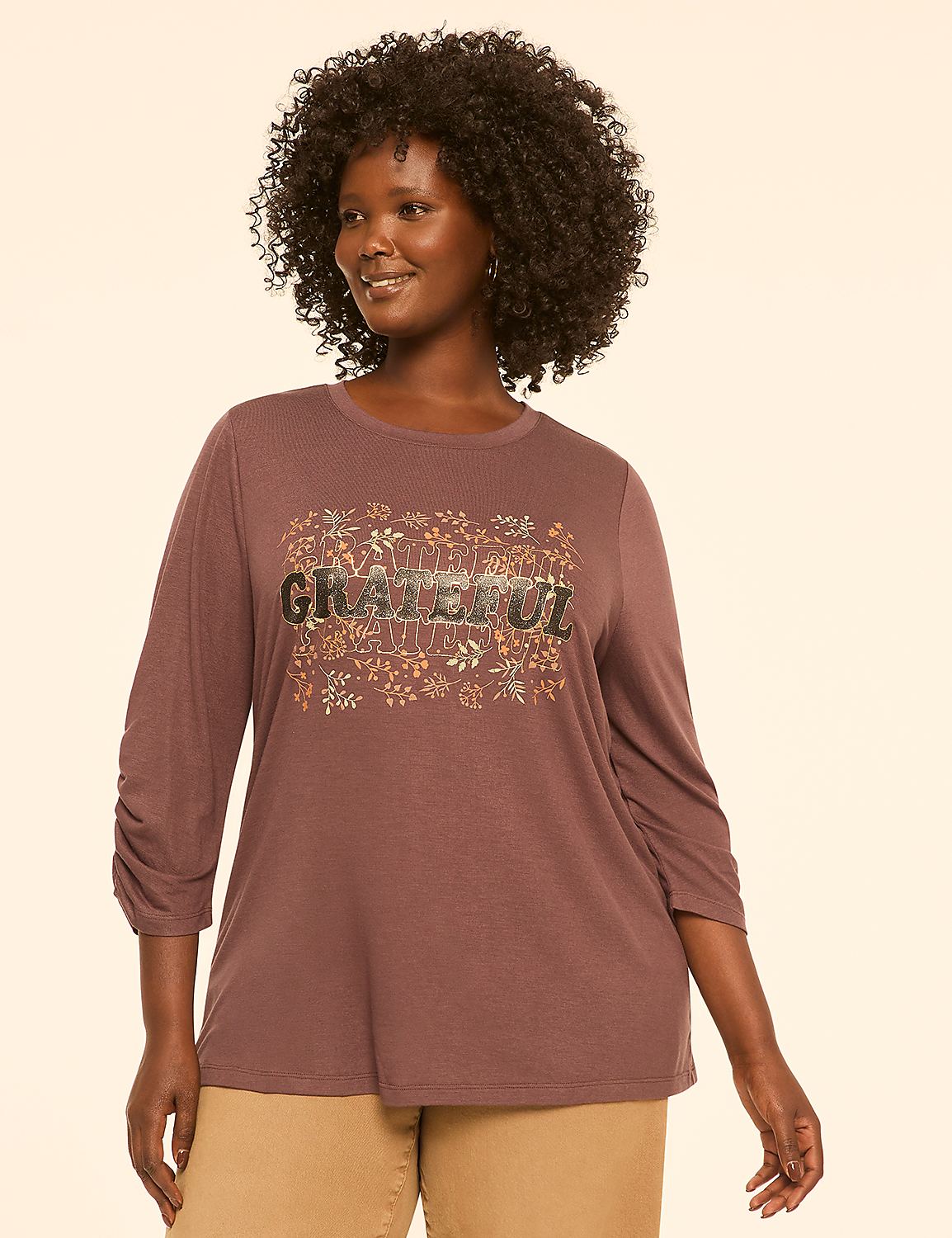 A woman in a mauve, ruched-sleeve top with "GRATEFUL" and floral embellishments stands against a plain, light tan background. Product Image 1