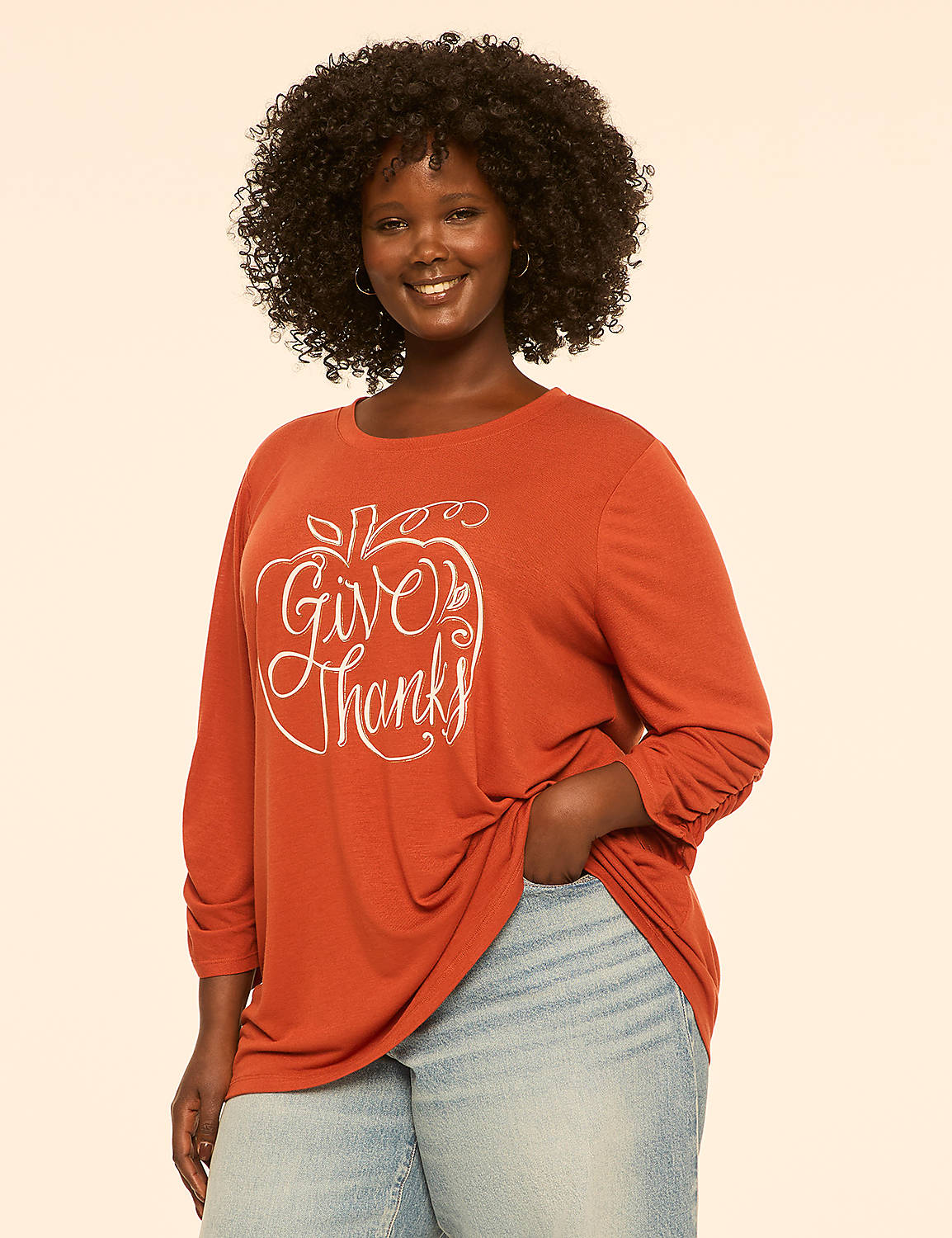 A smiling woman with curly dark hair wears an orange long-sleeved shirt with a "Give Thanks" apple graphic and light blue jeans against a plain cream background. Product Image 1