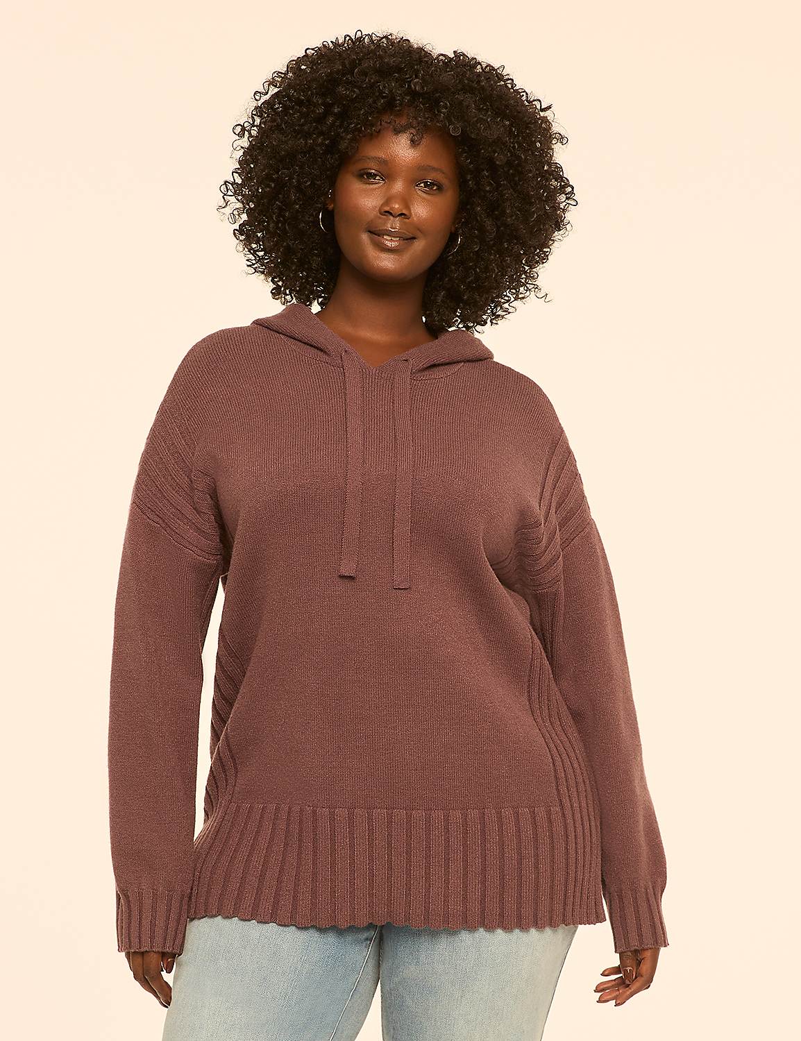 A woman with dark, curly hair smiles at the camera, wearing a mauve hooded sweater and light blue jeans against a plain, light peach background. Product Image 1