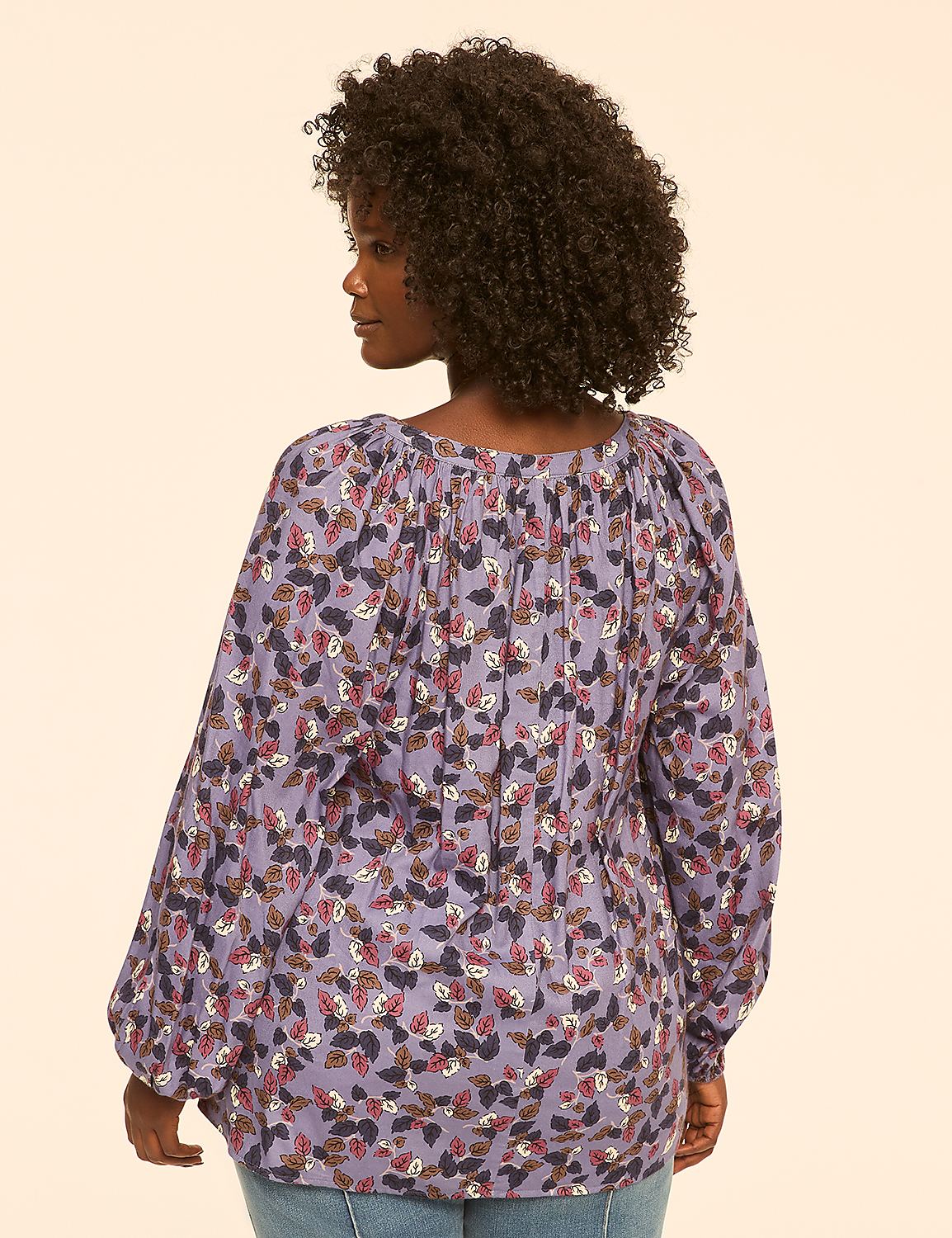A woman with curly dark hair wears a long-sleeved, purple, floral-patterned blouse and jeans, standing against a plain, light-colored background. Product Image 2