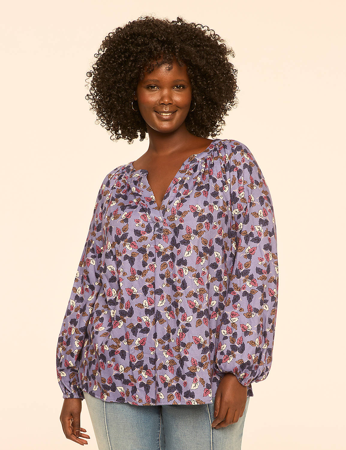 A smiling woman with curly dark hair wears a purple floral blouse and light blue jeans against a plain cream background. Product Image 1