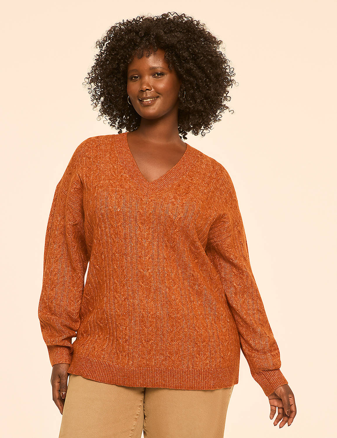 A woman with curly dark hair smiles in a textured orange v-neck sweater and tan pants against a plain beige background. Product Image 1