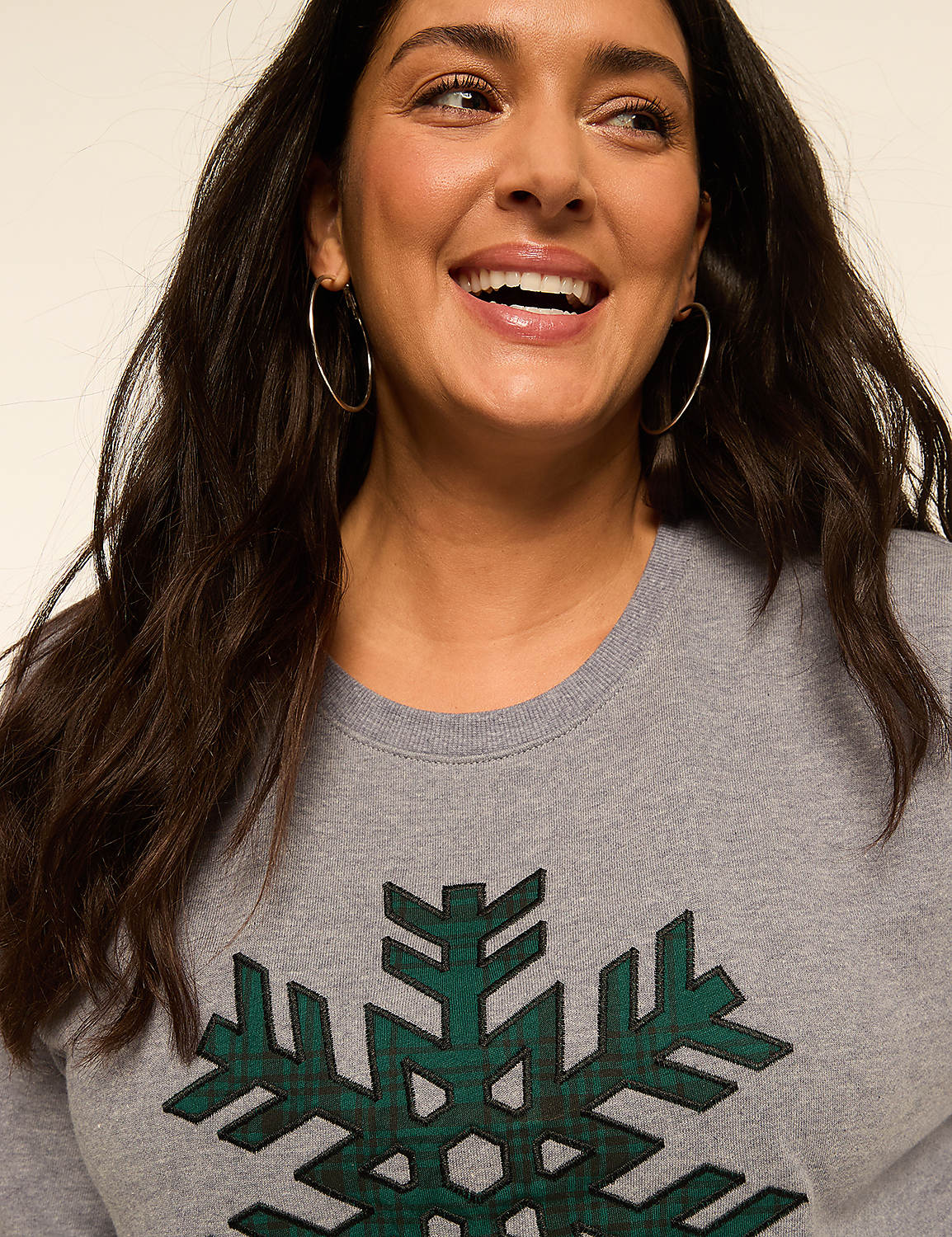 A smiling woman with dark hair and hoop earrings wears a grey sweatshirt with a green plaid snowflake design against a plain, light background. Product Image 4