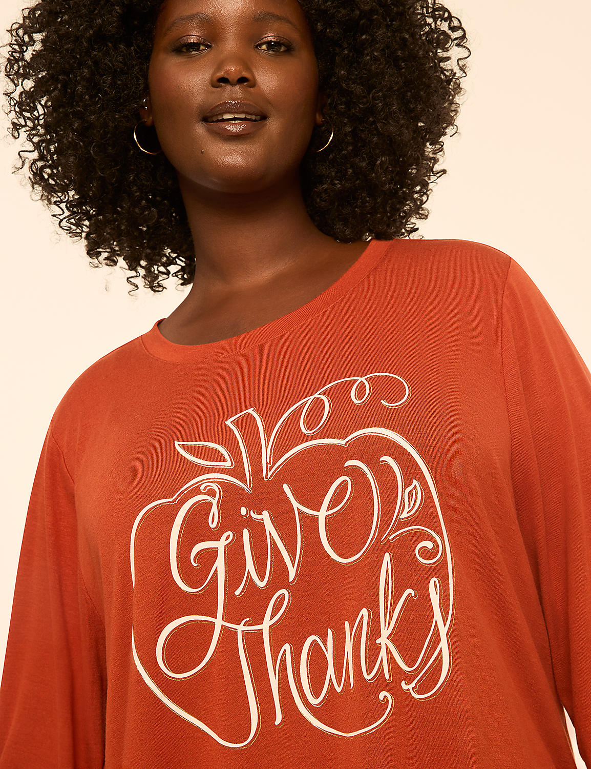 A woman with curly dark hair smiles as she wears an orange long-sleeved shirt with a white "Give Thanks" apple graphic, against a plain, light-colored background. Product Image 4
