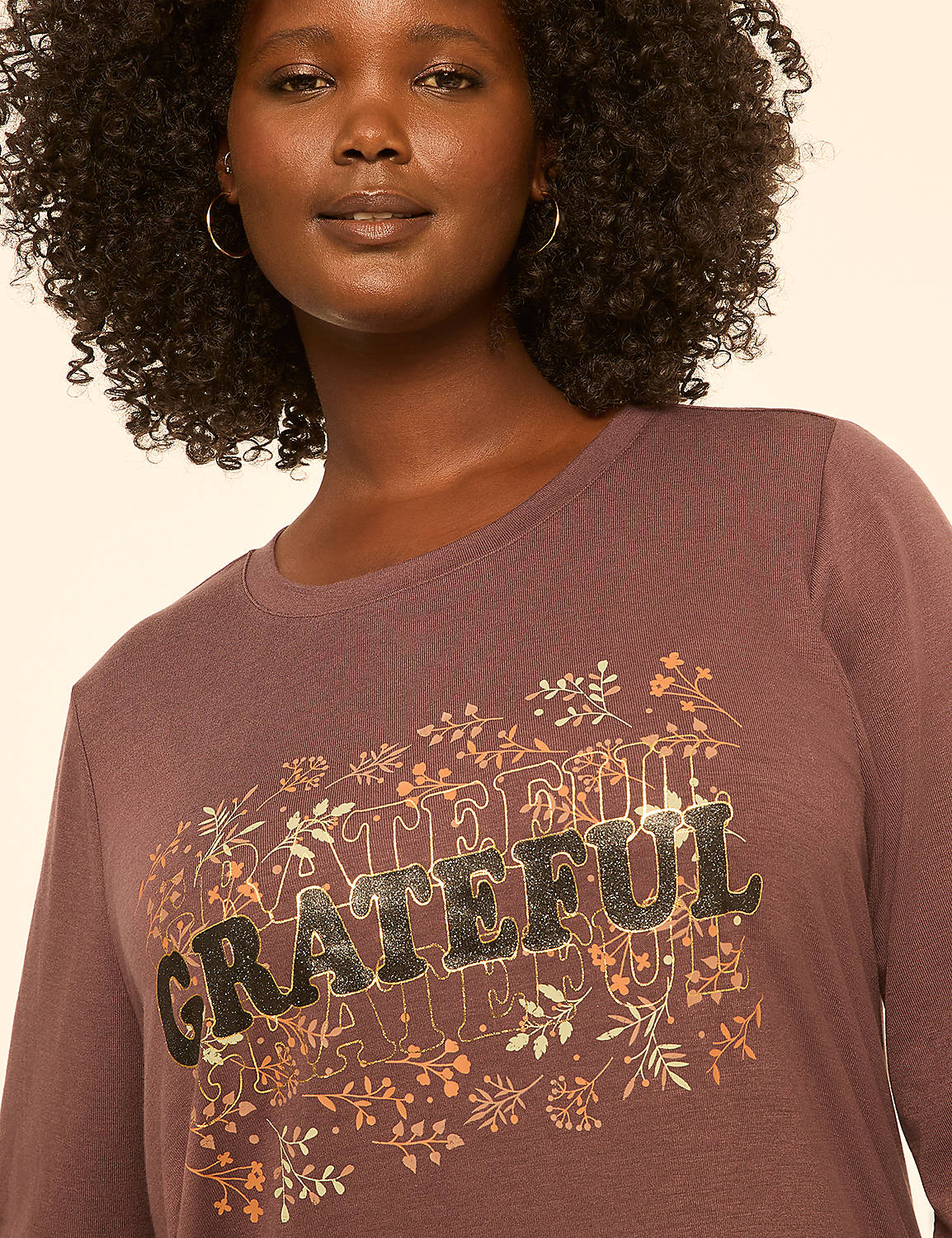 A woman with curly dark hair wears a mauve long-sleeved shirt with the word "GRATEFUL" in large, stylized letters surrounded by floral designs, against a plain, light-colored background. Product Image 4