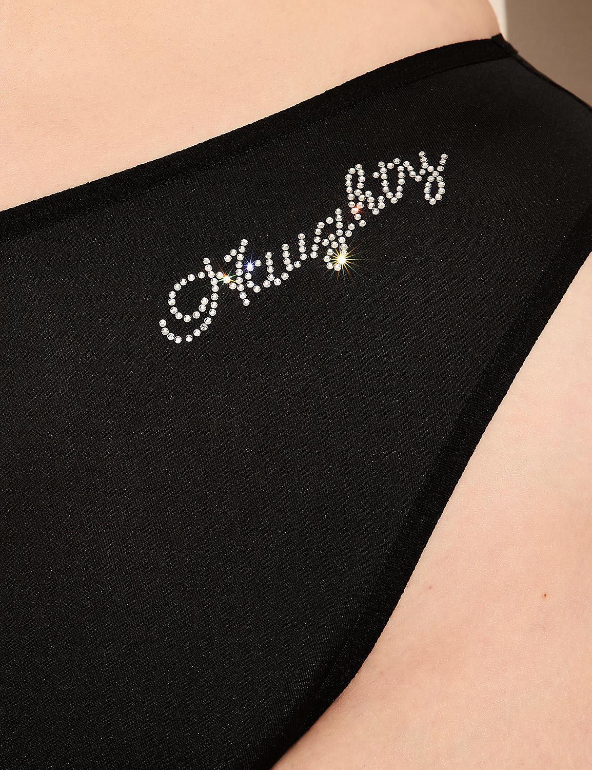 A black garment with the word "naughty" spelled out in rhinestones is shown against a blurred background of skin. Product Image 4