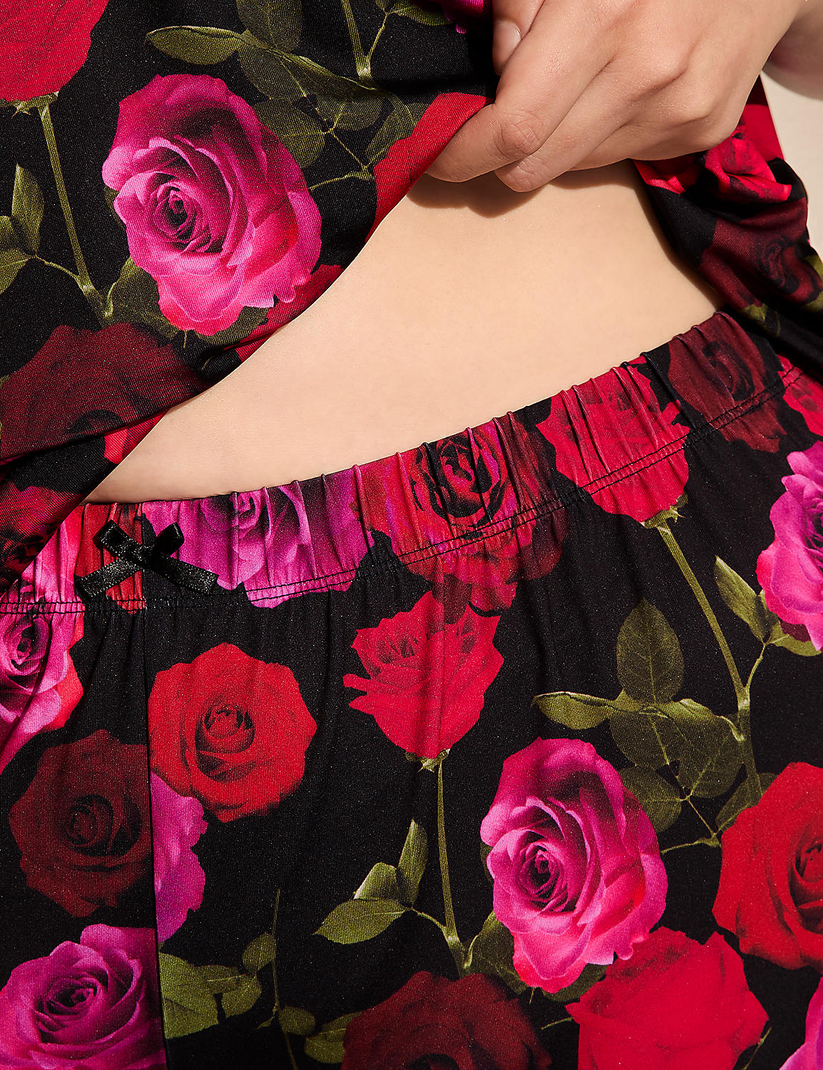A person's midsection is visible above floral patterned pajama bottoms featuring red and pink roses on a black background. Product Image 4