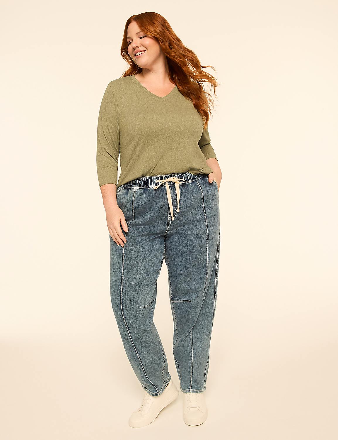 A smiling woman with red hair stands in the foreground wearing a green shirt and blue jeans against a plain, light-colored background. Product Image 3