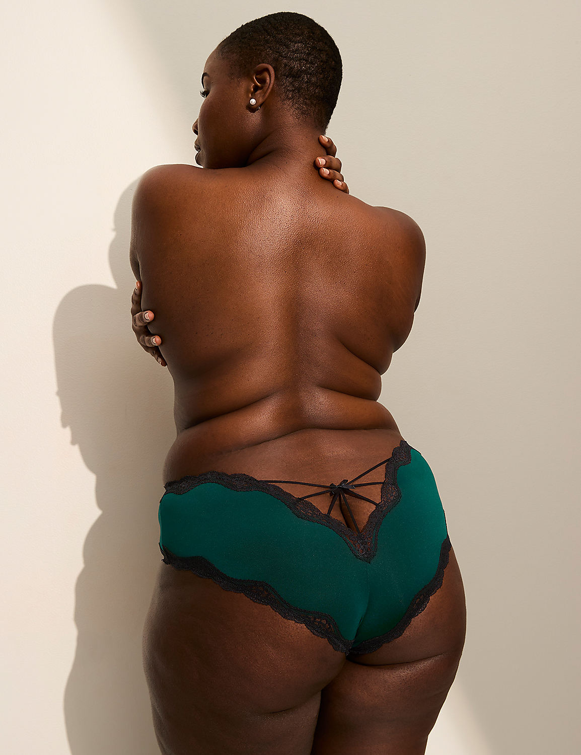 A woman with dark skin and short hair, wearing green lace-trimmed underwear, stands with her back to the viewer against a plain, light-colored wall with a shadow cast on it. Product Image 3