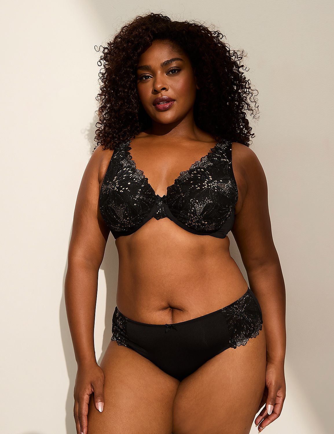 A woman with dark curly hair poses in a black lace bra and matching underwear against a plain, softly lit background. Product Image 4