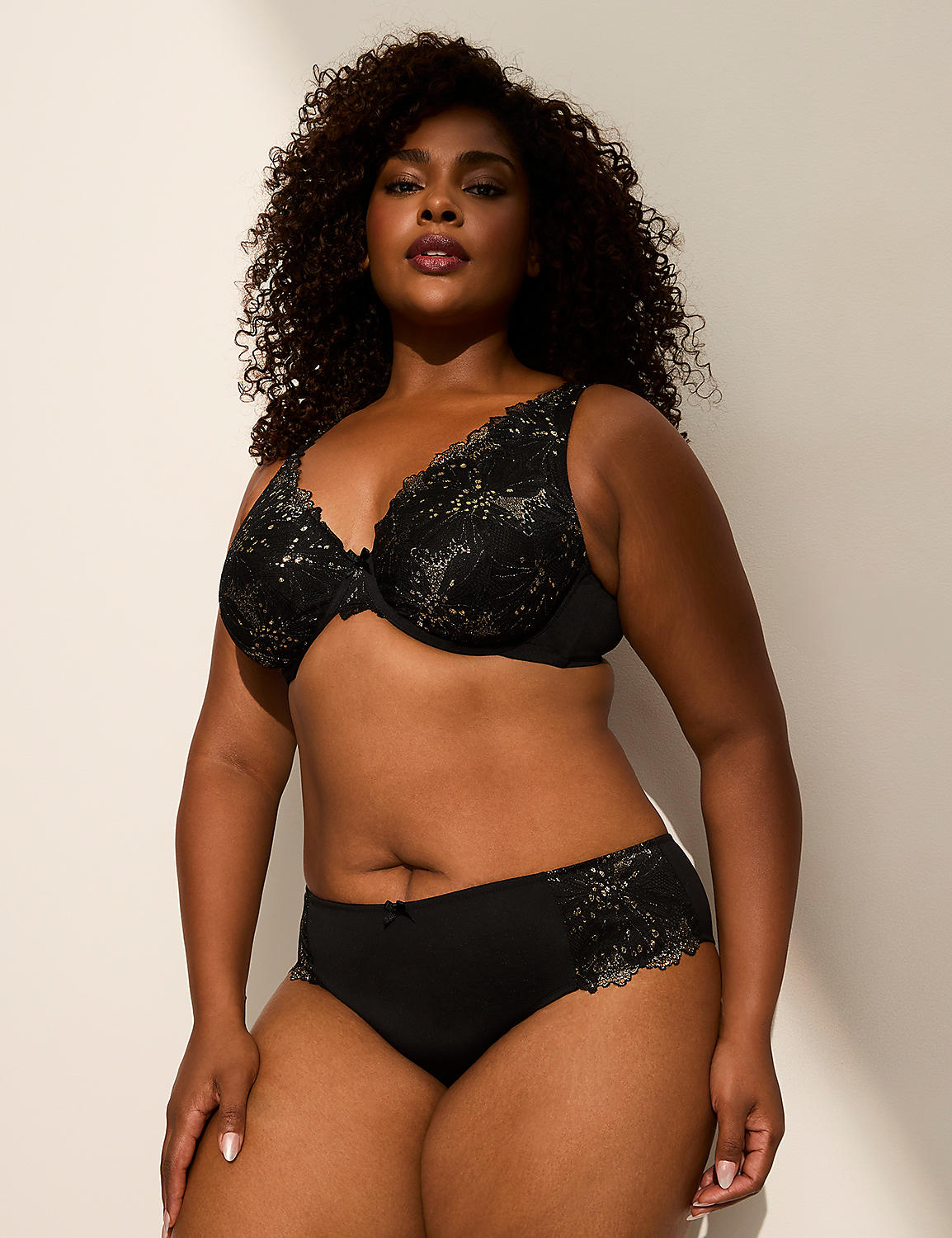 A woman with curly dark hair poses in a black lace bra and matching underwear against a plain, softly lit background. Product Image 3