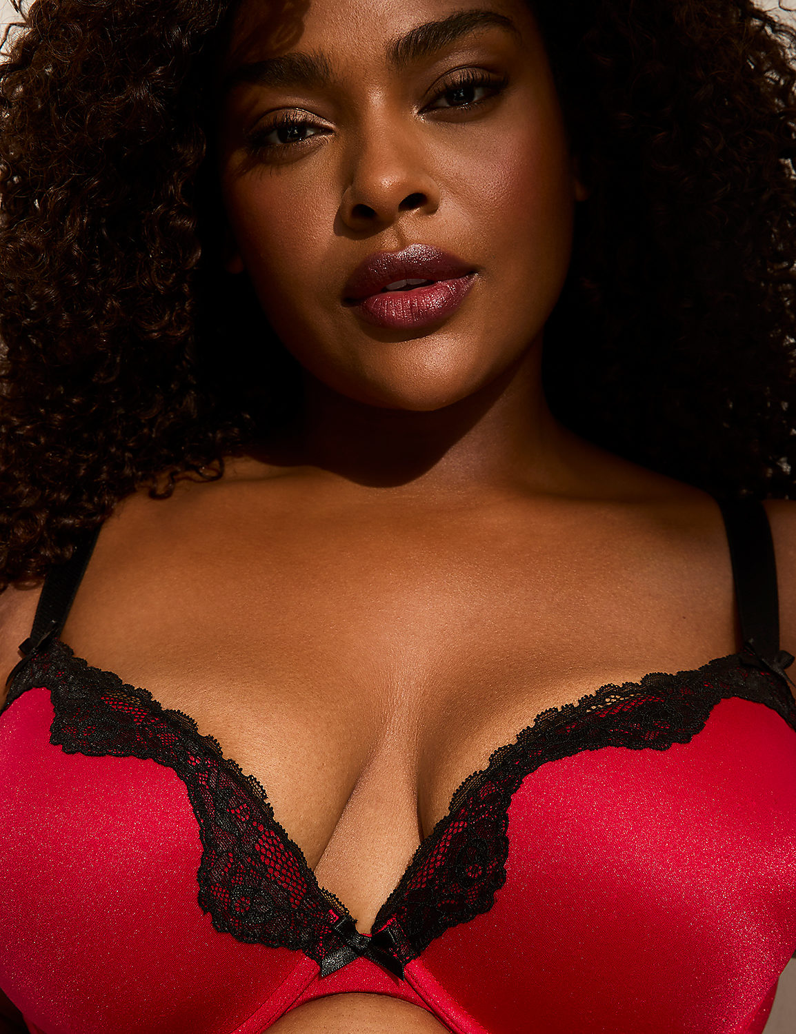 A woman with dark curly hair and dark lipstick is shown in the foreground wearing a red bra with black lace trim, against a plain background. Product Image 2