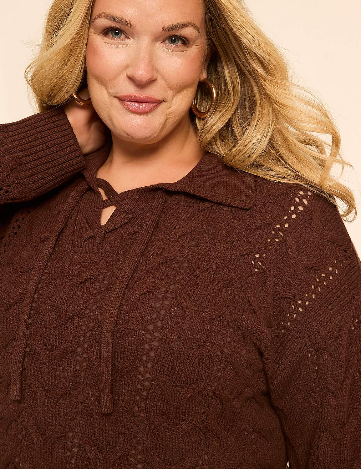 A woman with blonde hair and hoop earrings smiles as she wears a brown cable-knit sweater with a lace-up collar against a plain, light-colored background. Product Image 4
