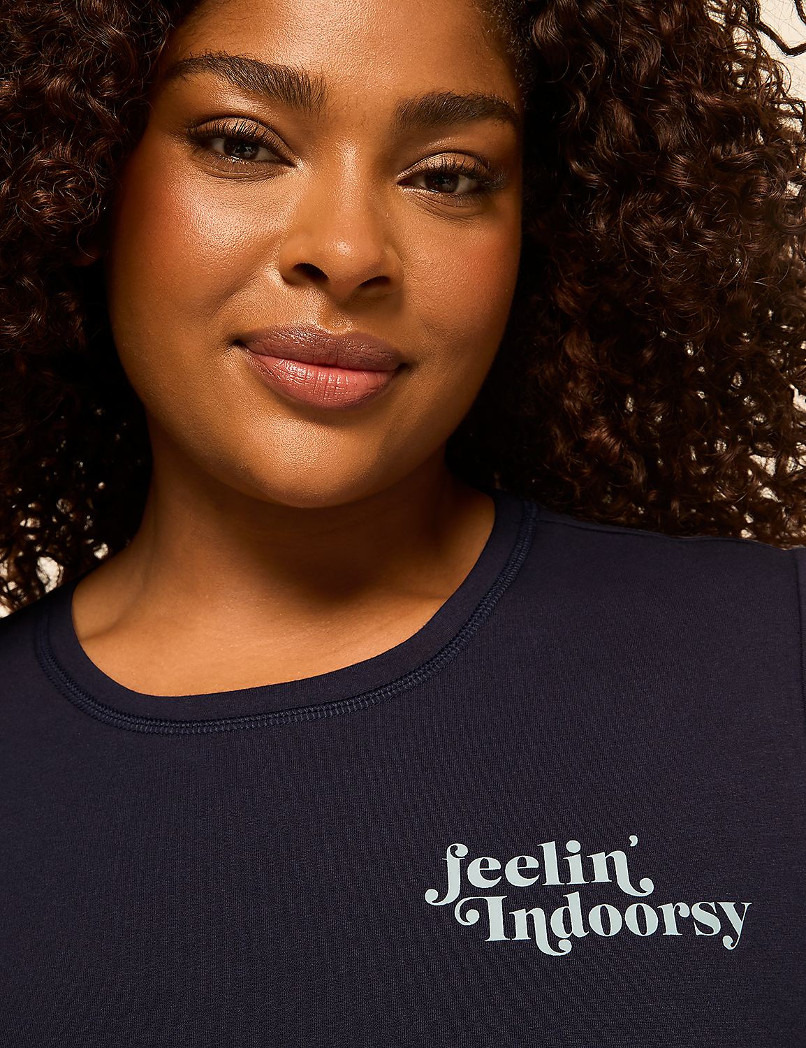 A woman with curly dark hair smiles gently in the foreground, wearing a dark blue t-shirt with the text "feelin' Indoorsy" in a light blue script, while the background is a dark, indistinct blur. Product Image 4