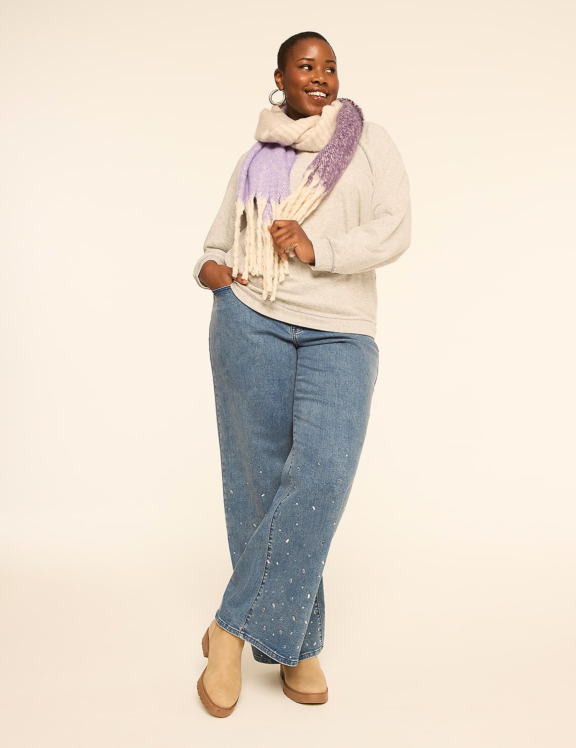 A woman in a light gray sweater, blue jeans adorned with rhinestones, and a purple and cream scarf stands against a plain, light-colored background. Product Image 3