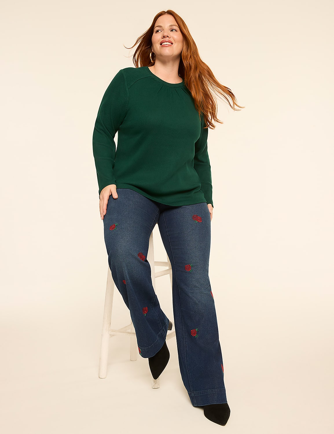 A woman with red hair sits on a white stool in the foreground, wearing a green long-sleeved shirt and wide-leg jeans with red embroidery, against a plain, light-colored background. Product Image 3