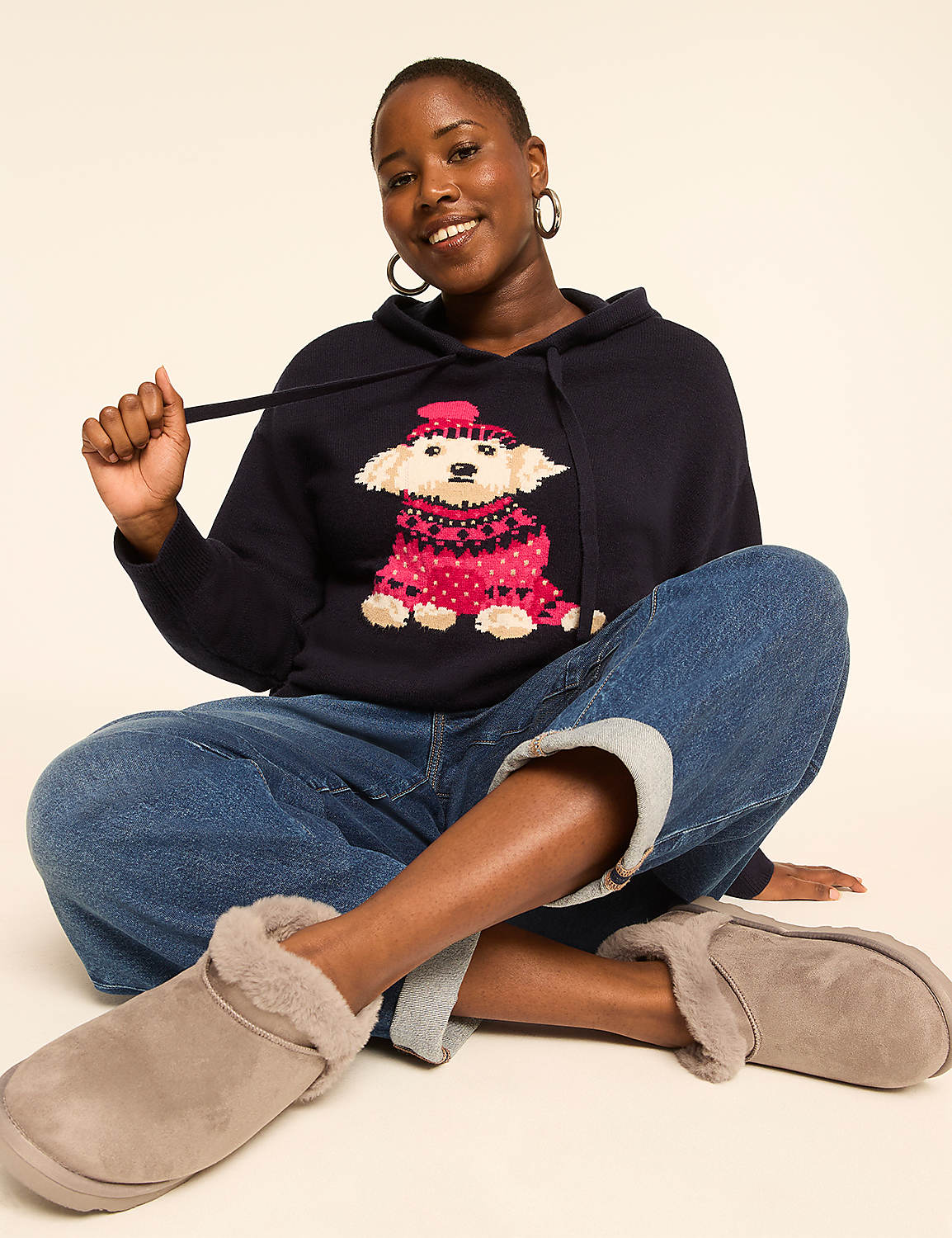 A woman in a dark blue hoodie with a dog design and rolled-up jeans sits cross-legged on a light-colored background, wearing fuzzy slippers. Product Image 3