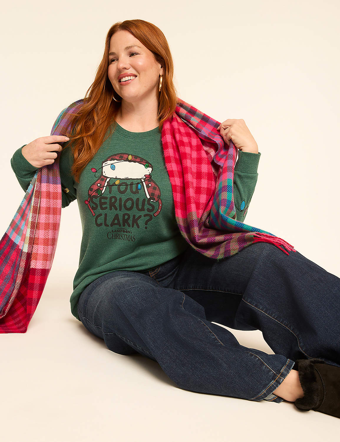 A smiling woman with red hair in a green sweater and jeans is wrapped in a colorful plaid scarf against a plain, light background. Product Image 3