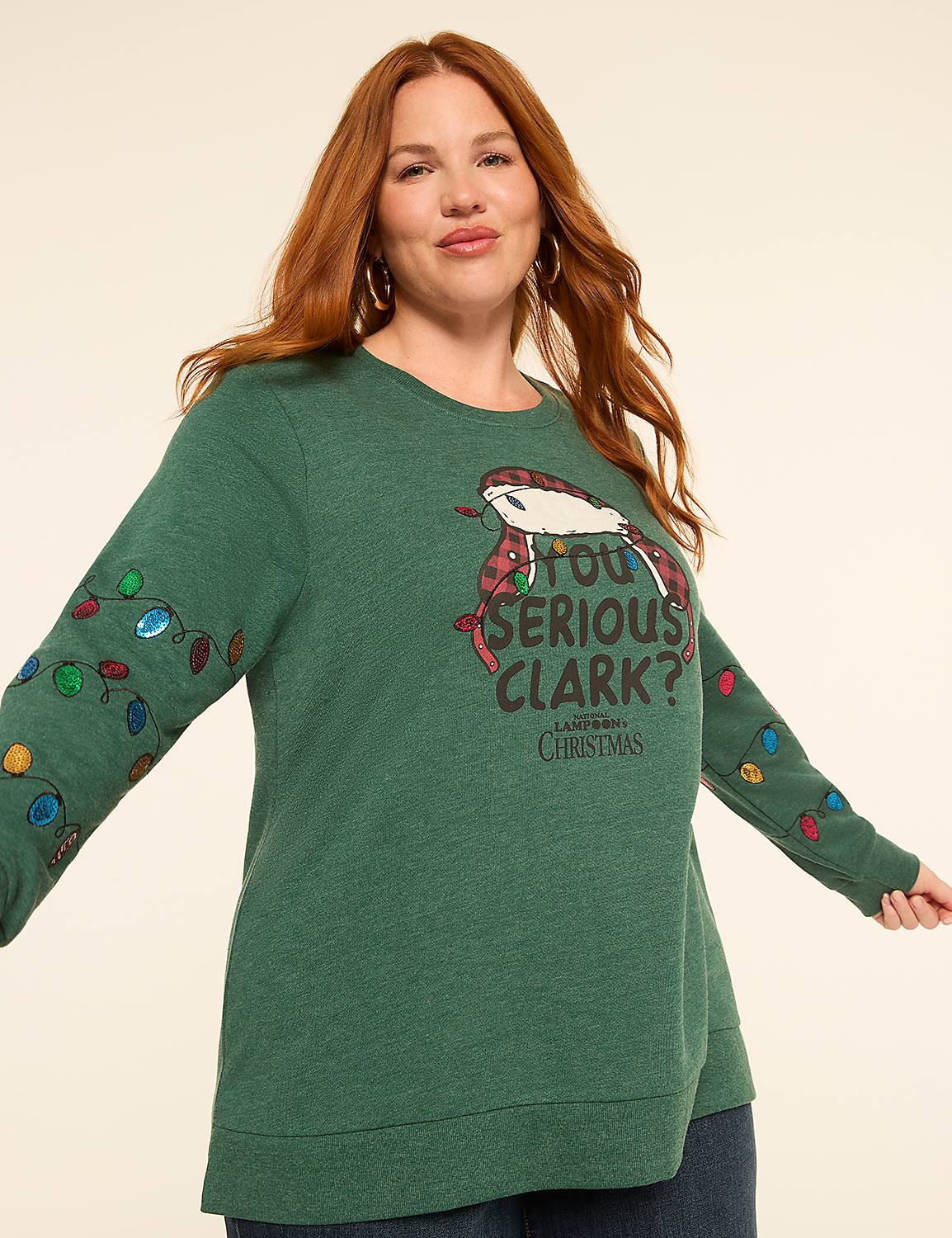 A woman with red hair smiles at the camera wearing a green Christmas sweater with a "You Serious, Clark?" graphic and colorful lights on the sleeves, against a plain, light-colored background. Product Image 1