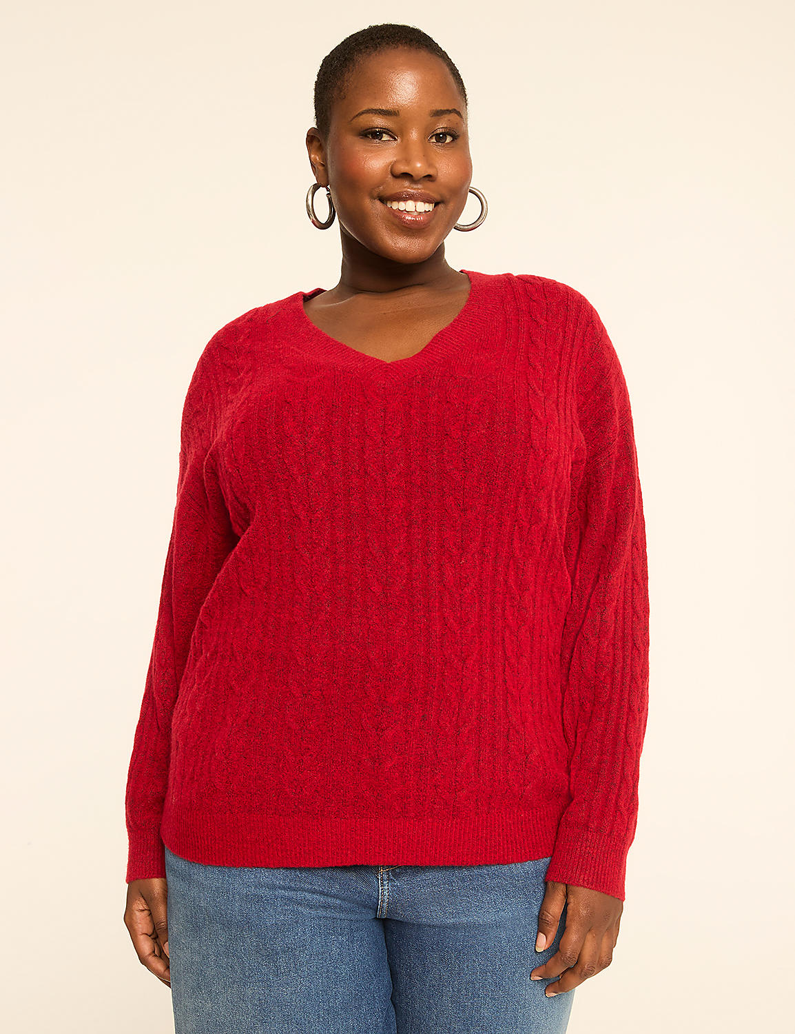 A smiling woman in a red cable-knit sweater and blue jeans stands against a plain, light-colored background. Product Image 1
