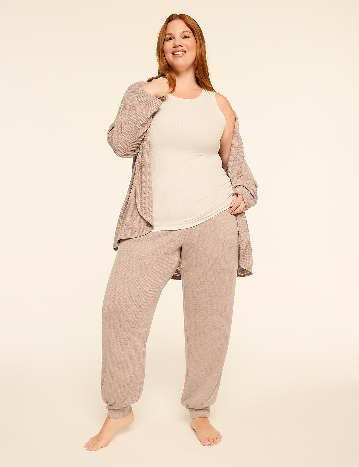 A smiling woman in a beige tank top, matching joggers, and a light brown cardigan stands barefoot against a plain, light beige background. Product Image 4