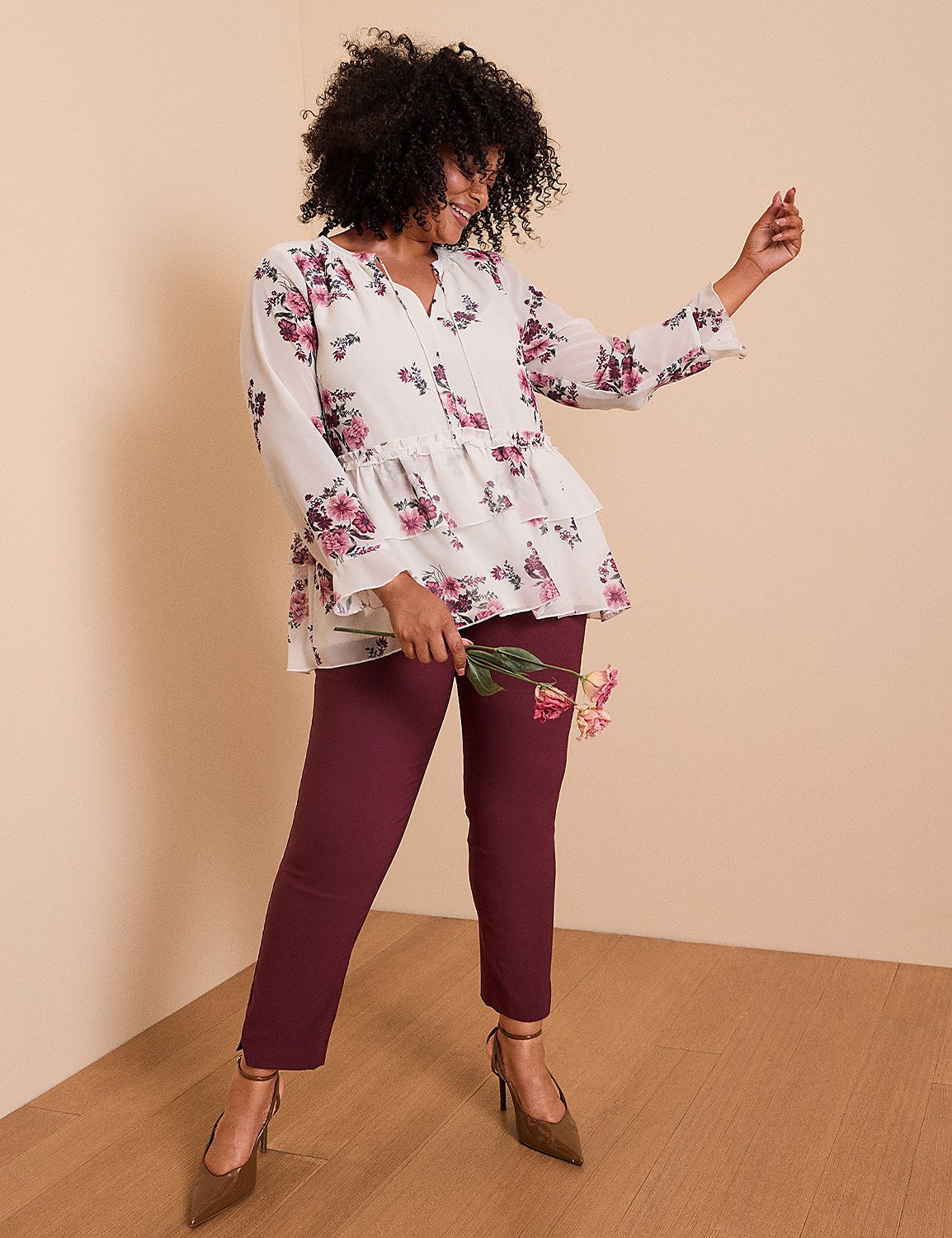 A woman in a floral blouse and maroon pants poses with a flower in her hand against a plain, light-colored wall. Product Image 1