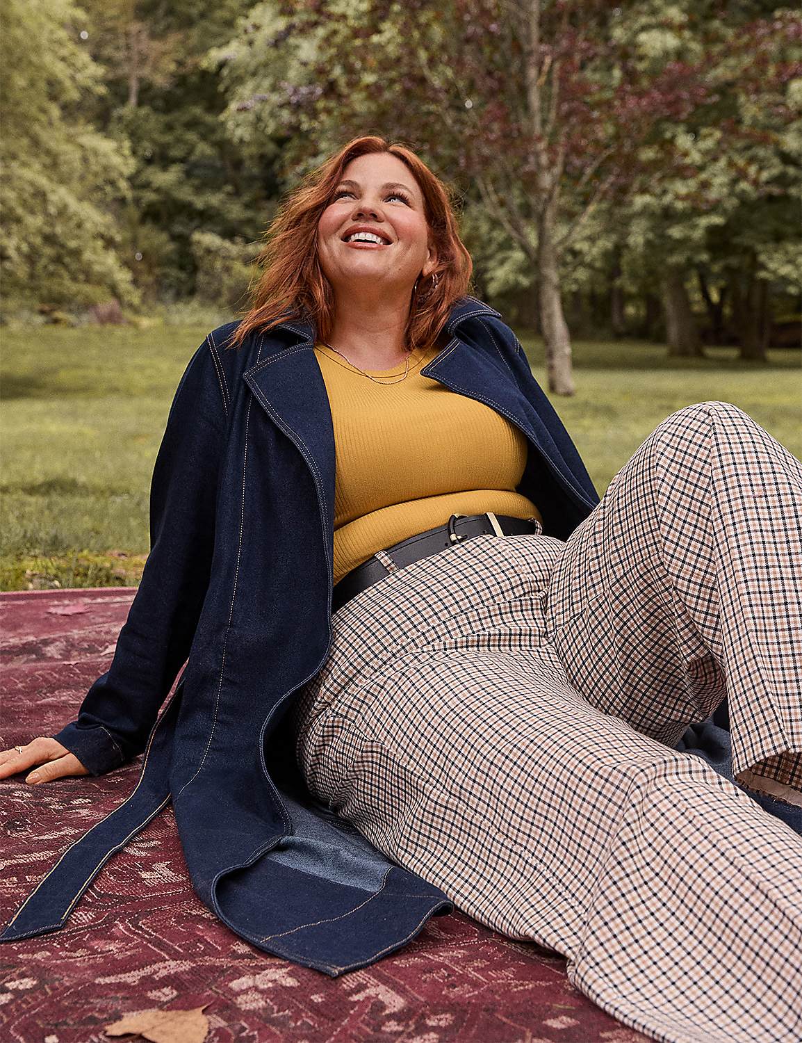 A smiling woman in a yellow top and plaid pants sits on a patterned rug in a park with trees in the background. Product Image 4