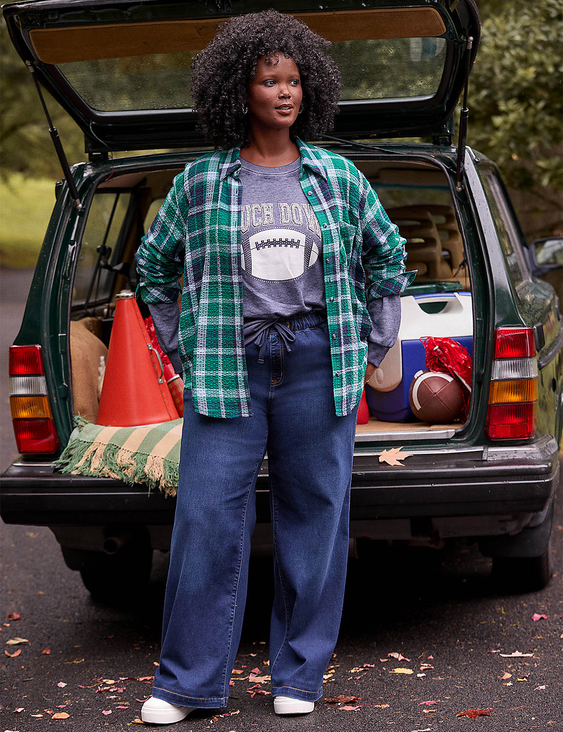 A woman in a green plaid shirt and jeans stands in front of an open car trunk filled with football-themed items, with trees in the background. Product Image 5