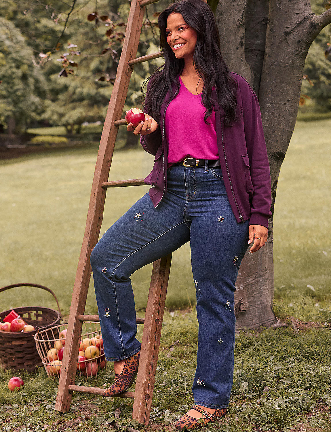 A woman in jeans and a cardigan stands by a ladder with apples in a basket in the foreground, while a tree and grassy yard form the background. Product Image 1