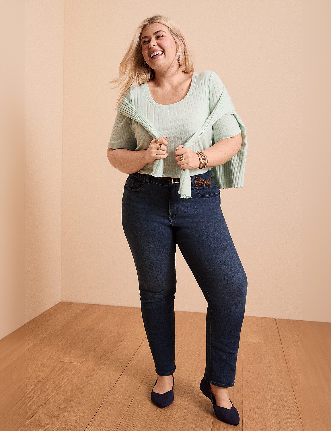 A smiling woman in a light green top and dark jeans stands on a wooden floor against a plain, light-colored wall. Product Image 4