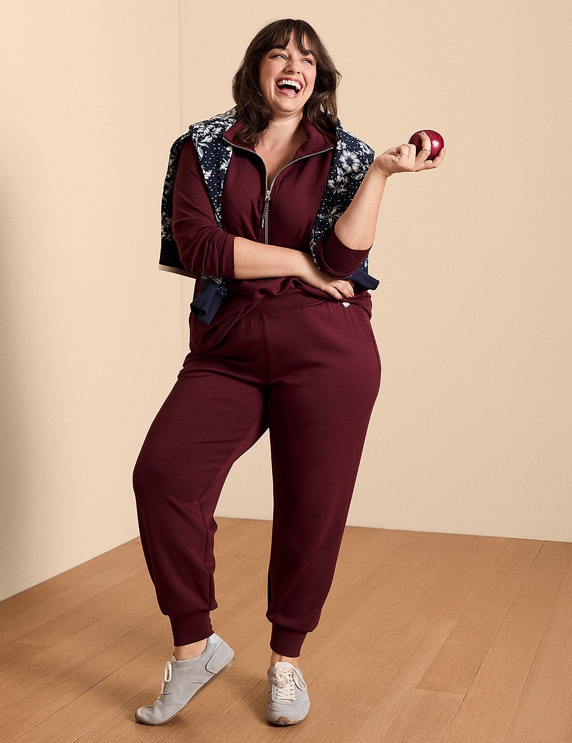 A woman in a maroon tracksuit and a patterned jacket laughs while holding an apple, against a plain beige background. Product Image 4