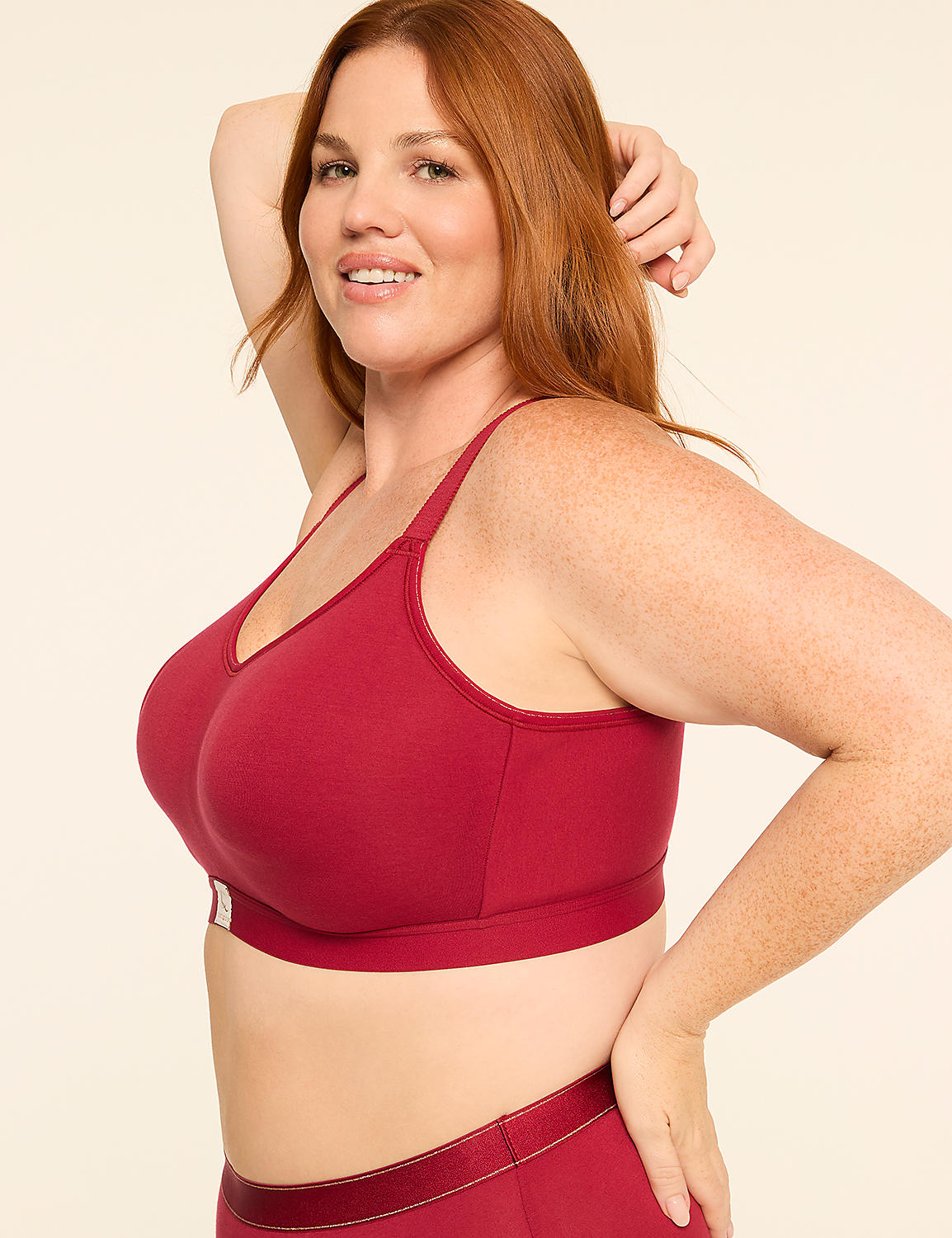 A woman with red hair and freckles poses in a maroon sports bra and matching bottoms against a plain cream background. Product Image 3