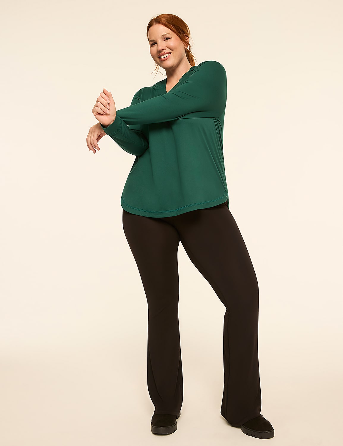 A woman in a green top and black pants stretches her arms in front of a plain, light-colored background. Product Image 3