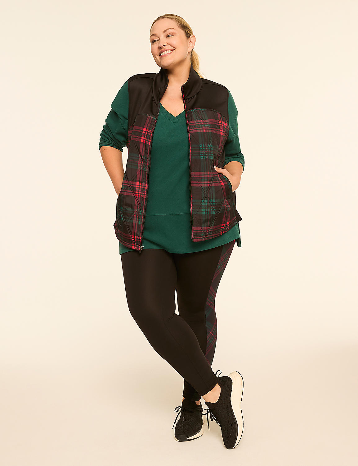 A smiling woman in a green top, plaid vest, and black leggings stands against a plain, light-colored background. Product Image 3