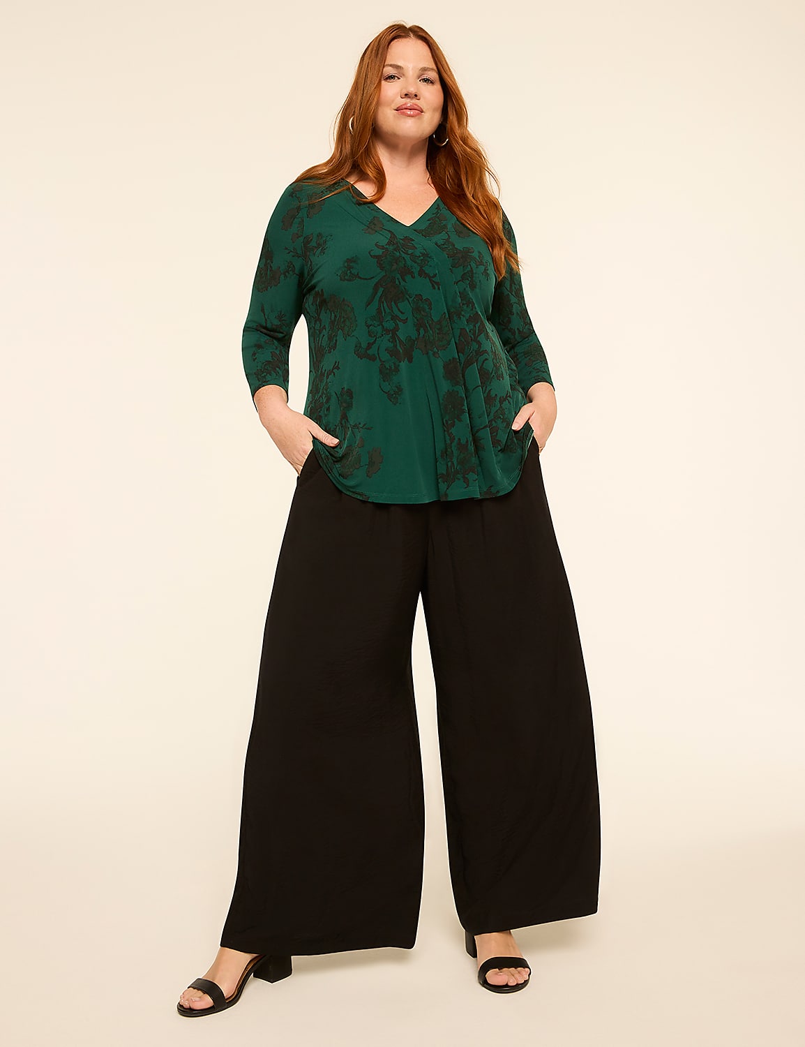 A woman with auburn hair stands with her hands in the pockets of her wide-leg black pants and a green floral-print top against a plain, light-colored background. Product Image 3