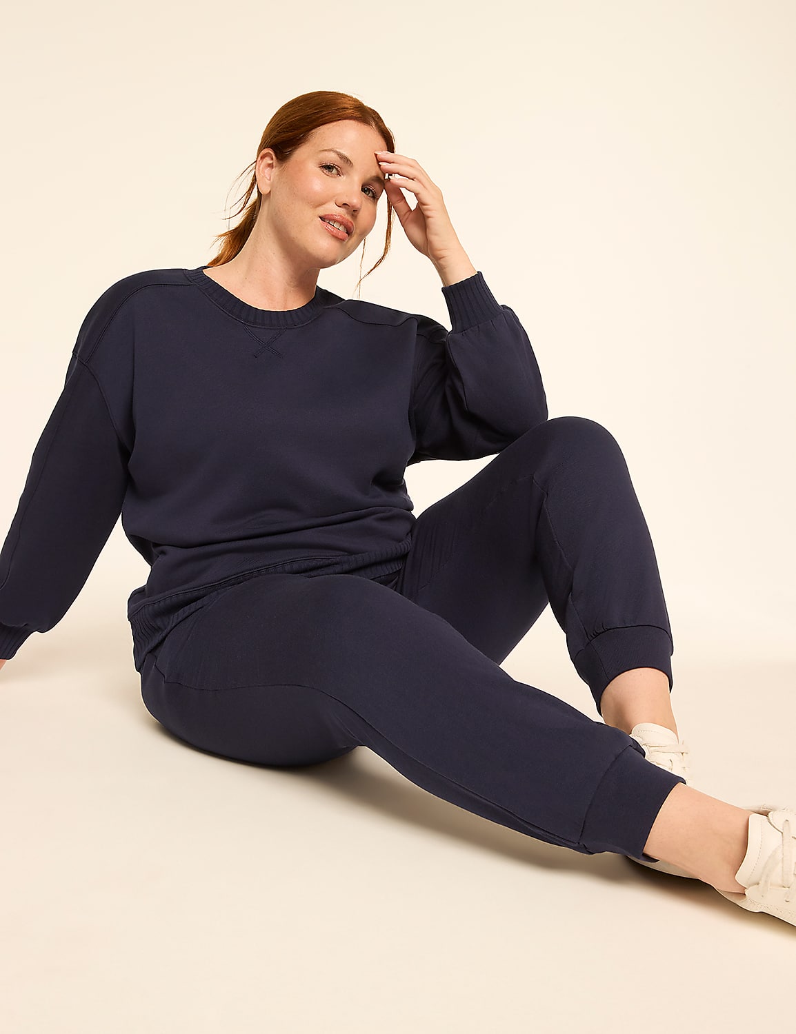 A woman in a navy blue tracksuit sits on a light beige background, with her hand resting on her head. Product Image 3