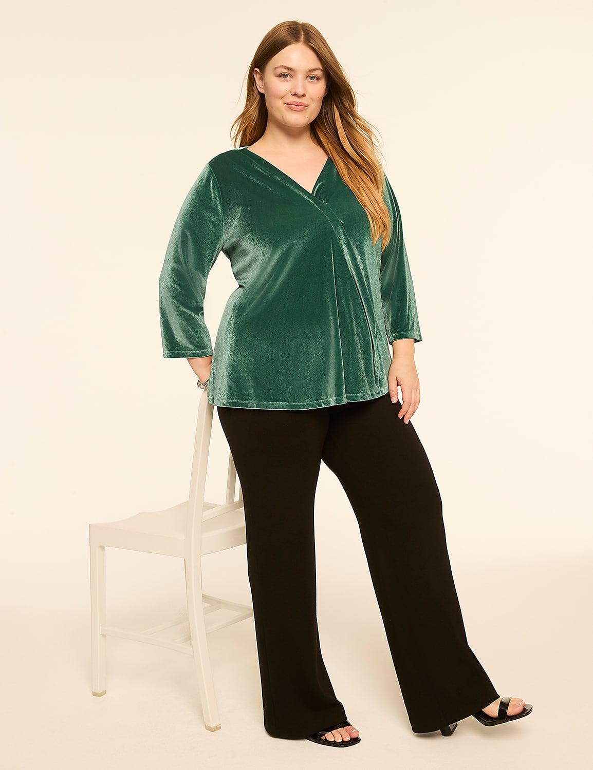 A woman in a green velvet top and black pants poses on a white chair against a cream-colored background. Product Image 3