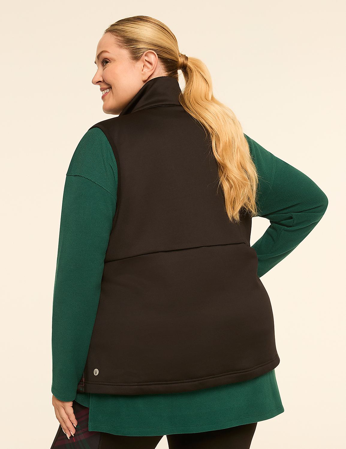 A woman with blonde hair pulled into a ponytail, wearing a black vest over a green long-sleeved shirt and plaid leggings, smiles over her shoulder against a plain, light-colored background. Product Image 2