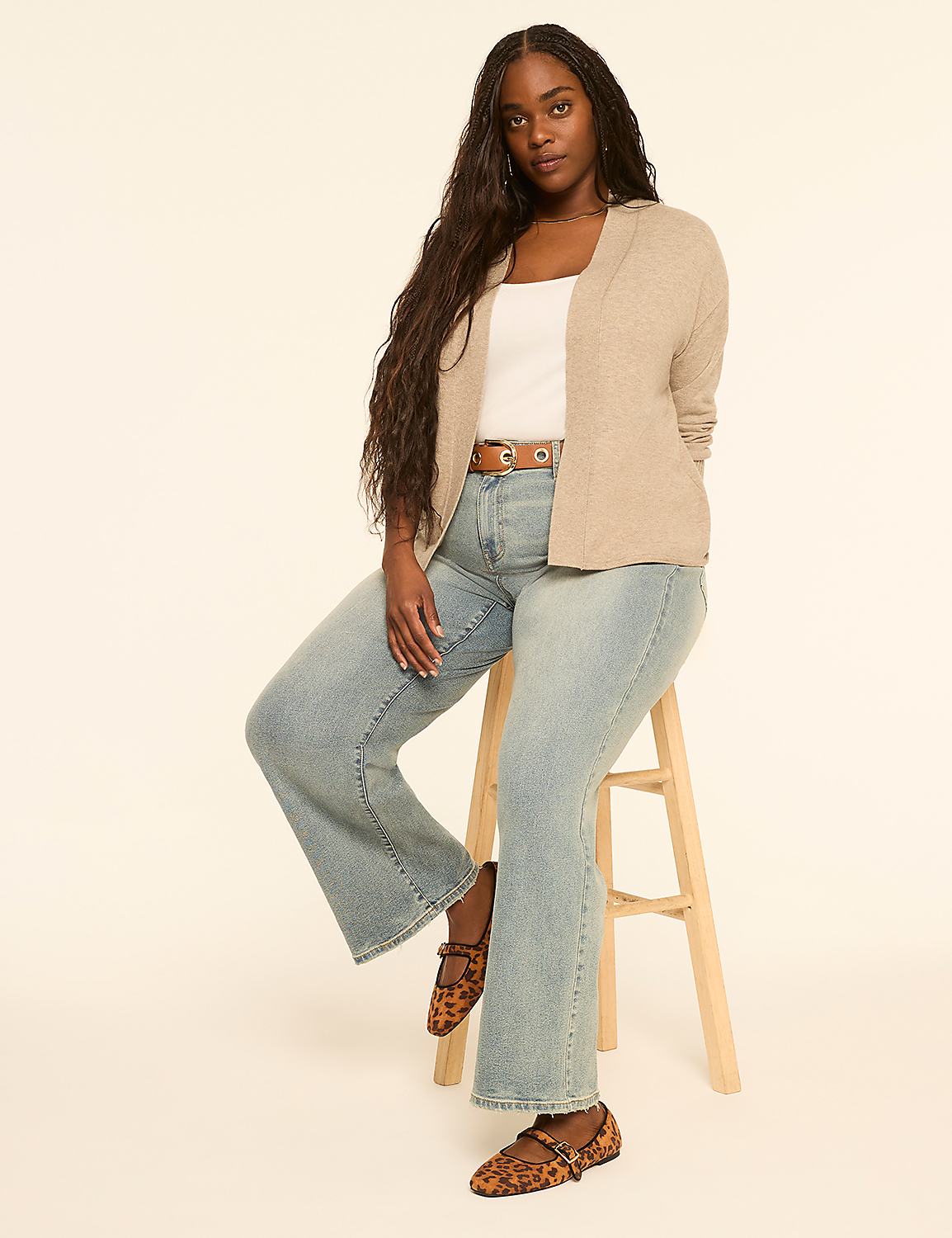 A woman with long, dark braided hair sits on a stool in the foreground, wearing a light-colored cardigan, white top, and light-wash flared jeans, against a plain, light-colored background. Product Image 3