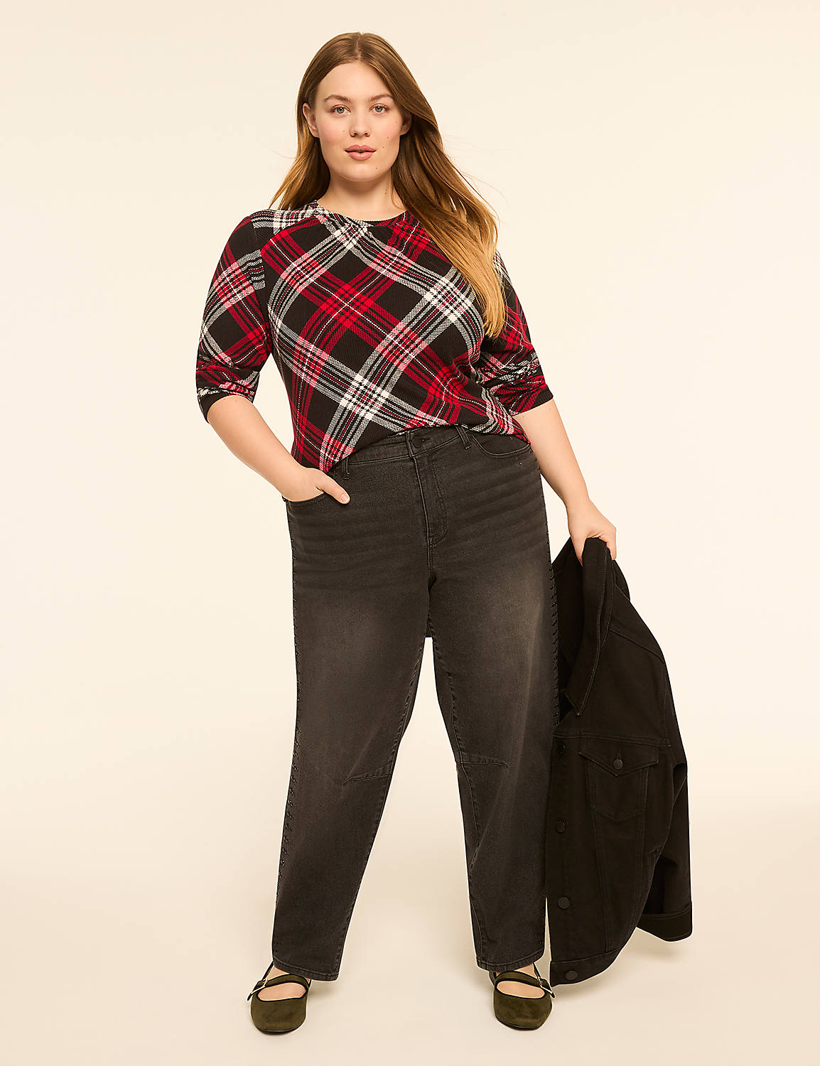 A woman in a plaid top and dark jeans stands against a plain, light-colored background, holding a dark jacket. Product Image 3