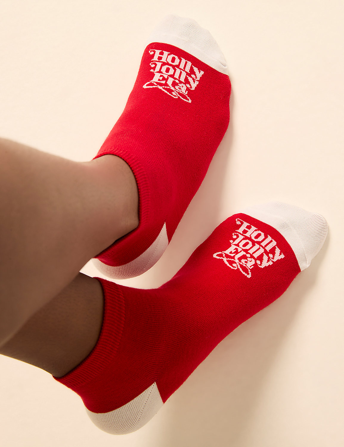 Red ankle socks with white toes and "Holly Jolly" logos are worn on a person's legs against a plain, light background. Product Image 1