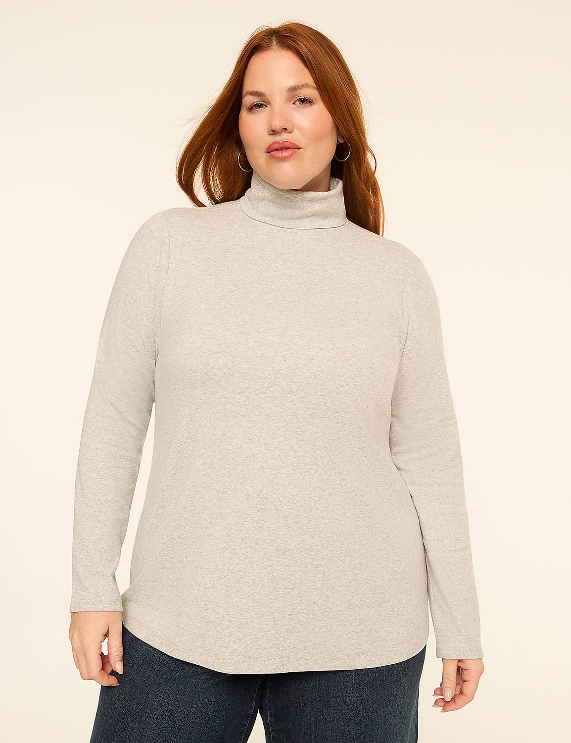 A woman with red hair and fair skin, wearing a light gray turtleneck sweater and dark blue jeans, stands against a plain, light beige background. Product Image 1